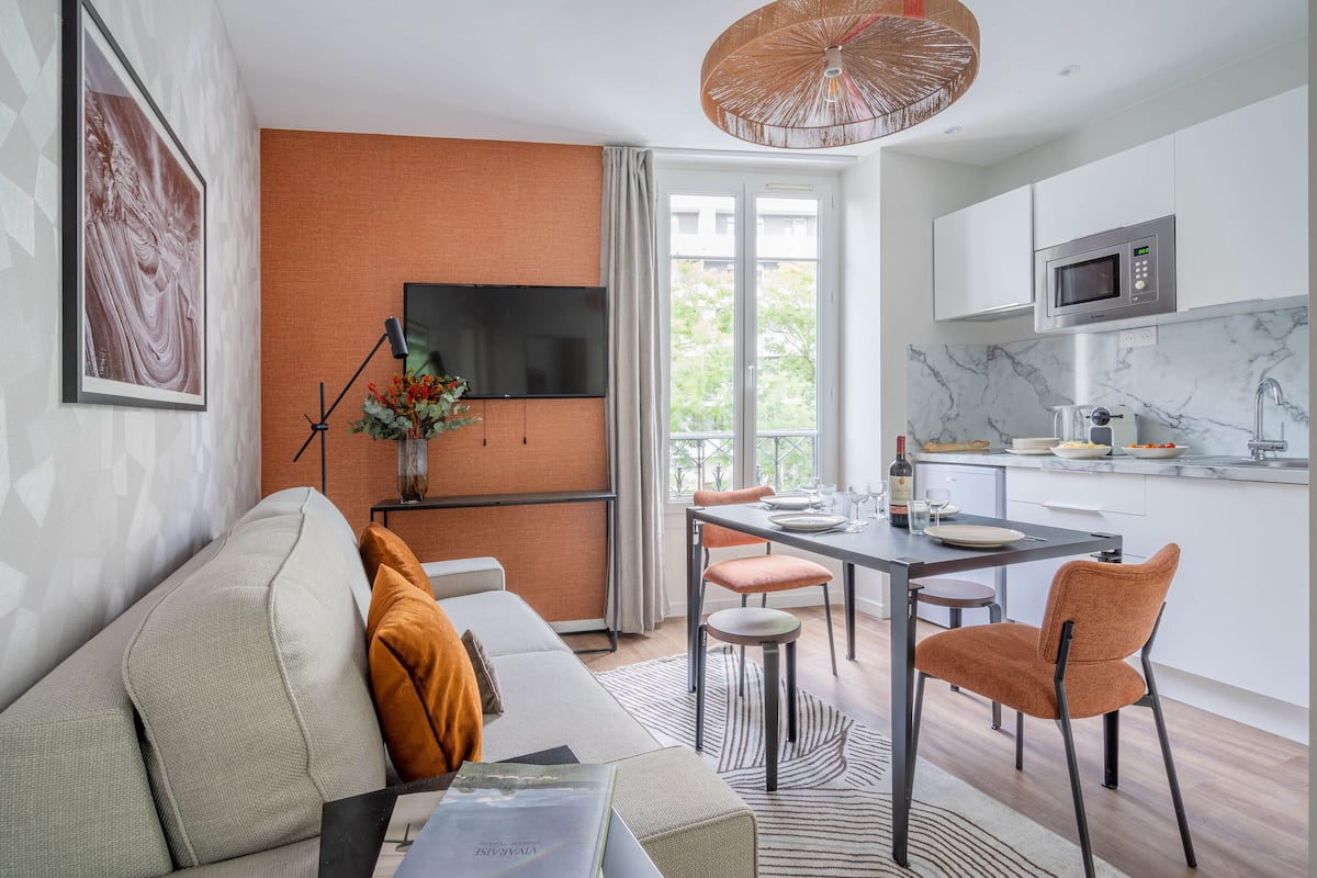 A modern living room with a sofa, a dining table with chairs, a TV mounted on the wall, and a kitchen area with a microwave.
