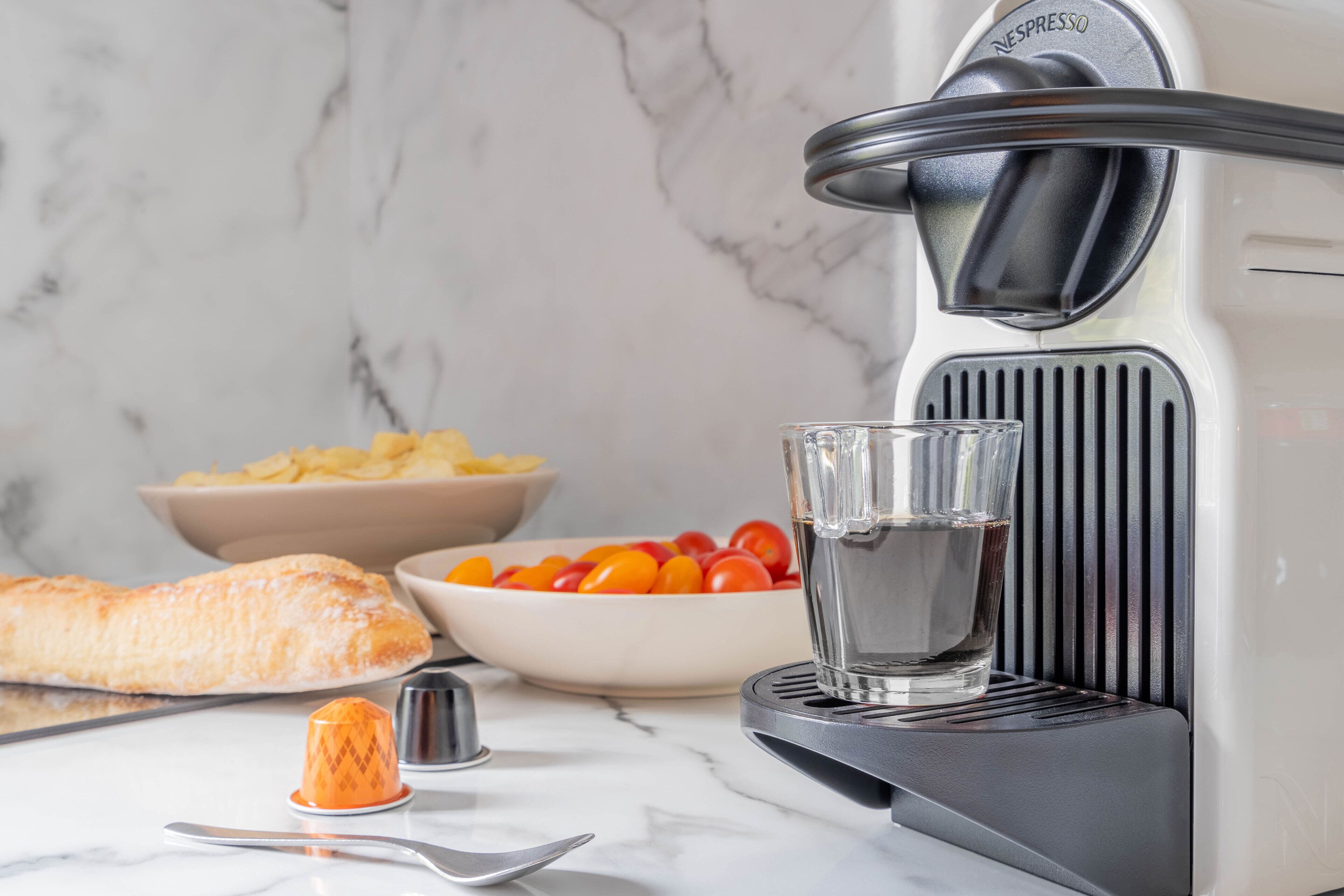 Een keuken met een koffieapparaat, een kom tomaten, een glas water en een croissant op het aanrecht.