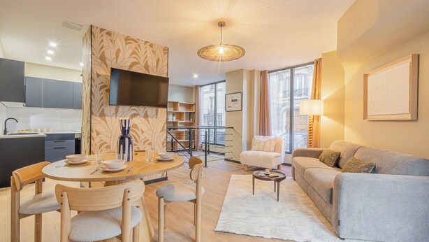 A modern living room with a sofa, dining table, and chairs. There is a kitchen area with cabinets and a TV. A staircase is visible in the background. - Residence Le Belleville (Paris)
