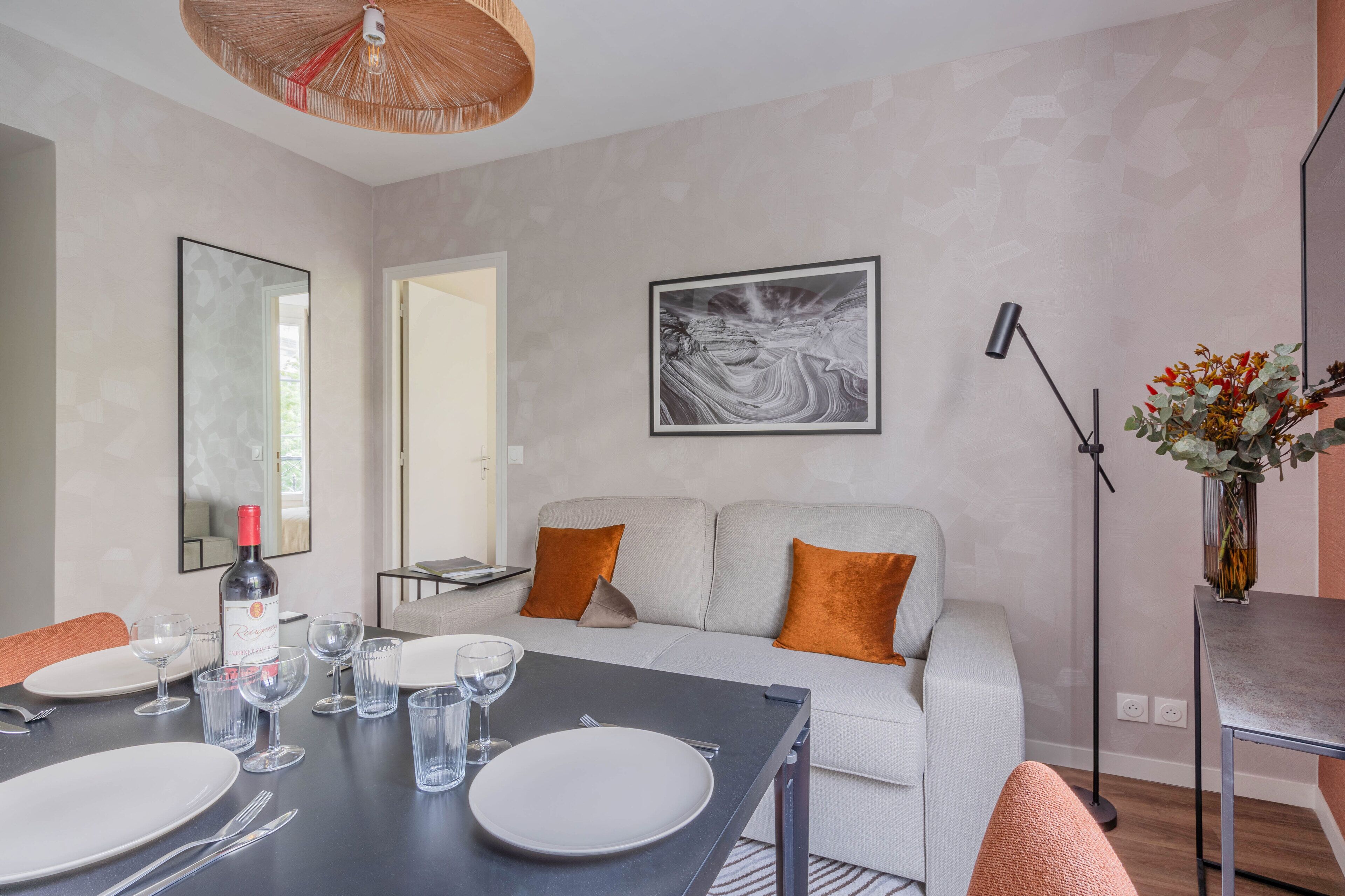 A modern living room with a dining table set for four, a sofa with orange cushions, a large abstract painting, and a floor lamp.
