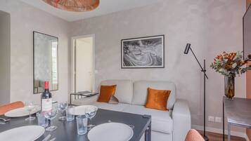 Una sala de estar moderna con una mesa de comedor para cuatro, un sofá con cojines naranja, un cuadro abstracto grande y una lámpara de pie.