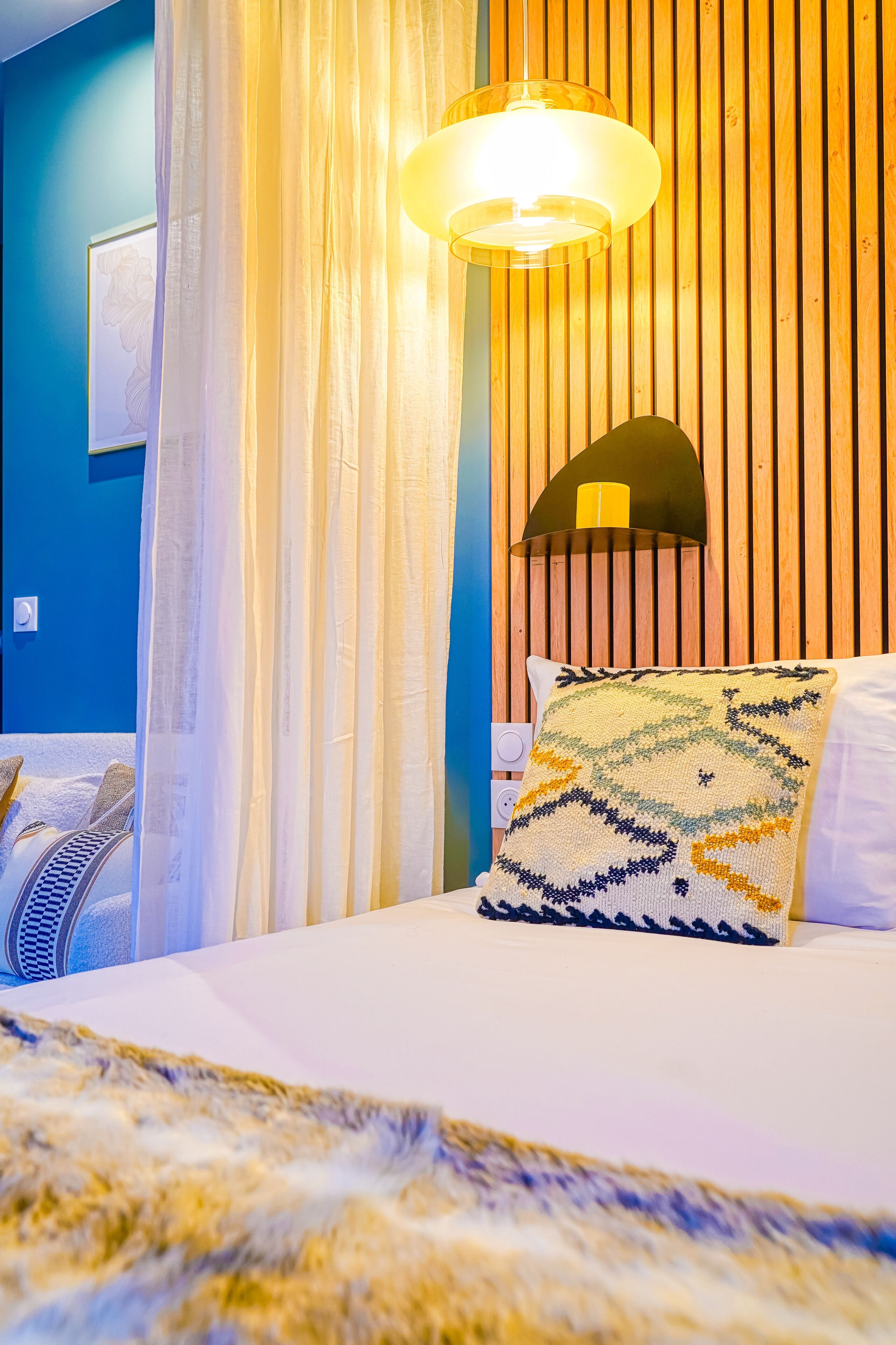 A hotel room with a bed, a bedside lamp, a wall-mounted fan, and a curtain.