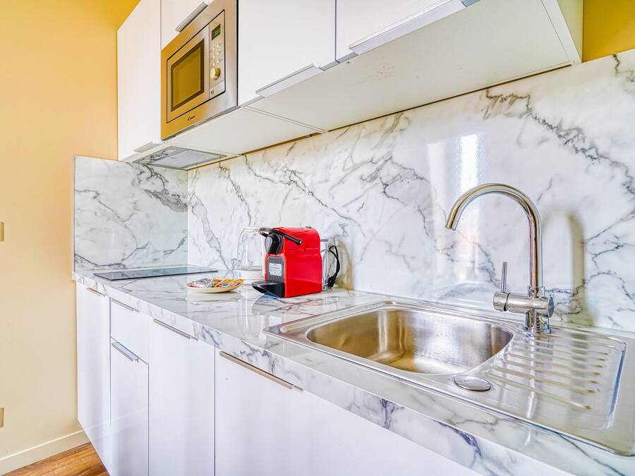 A kitchen with white cabinets, a marble backsplash, a stainless steel sink, and a red coffee maker.