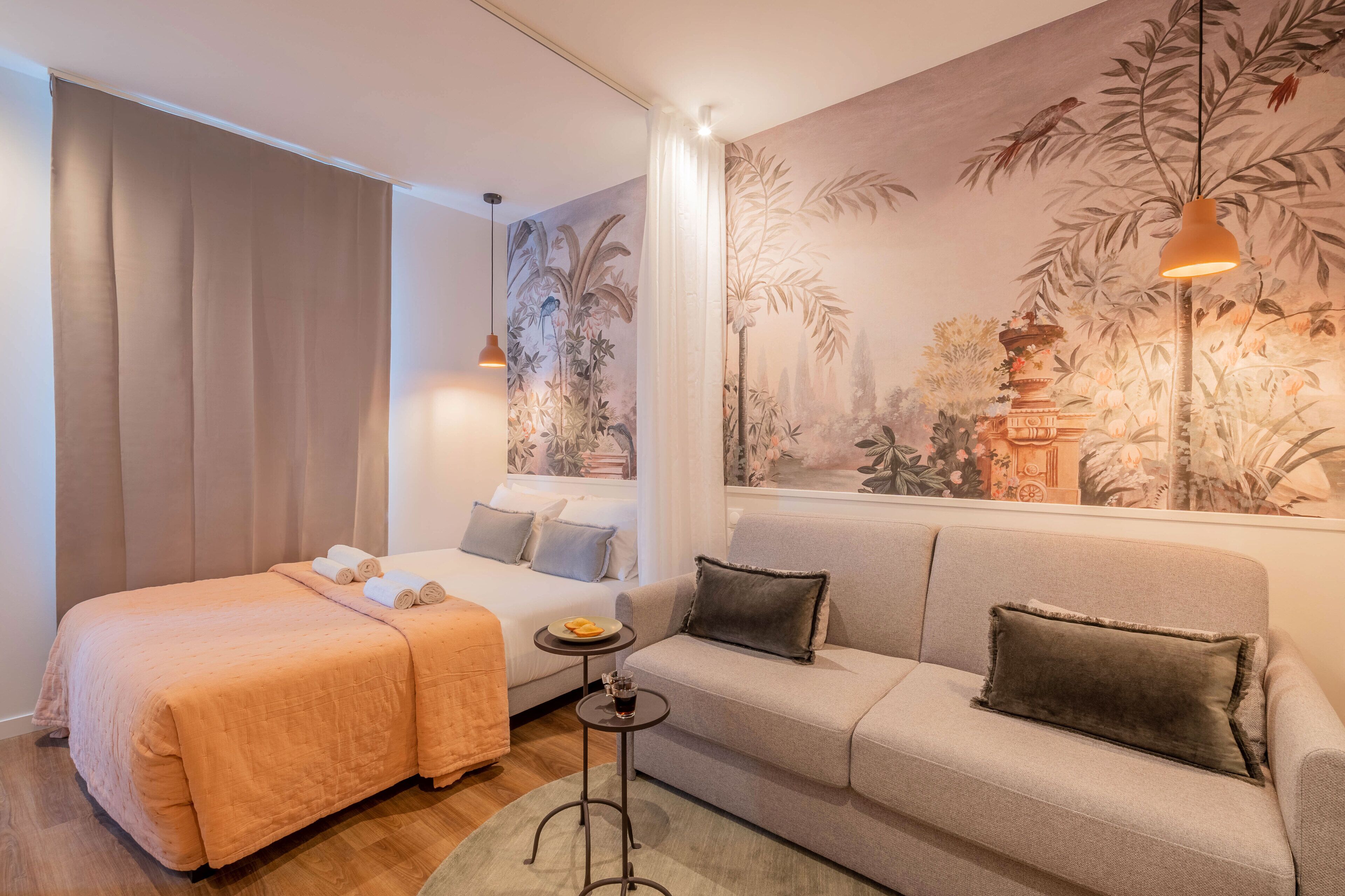 A hotel room with a bed, a sofa, a small table with a drink, and a mural on the wall.