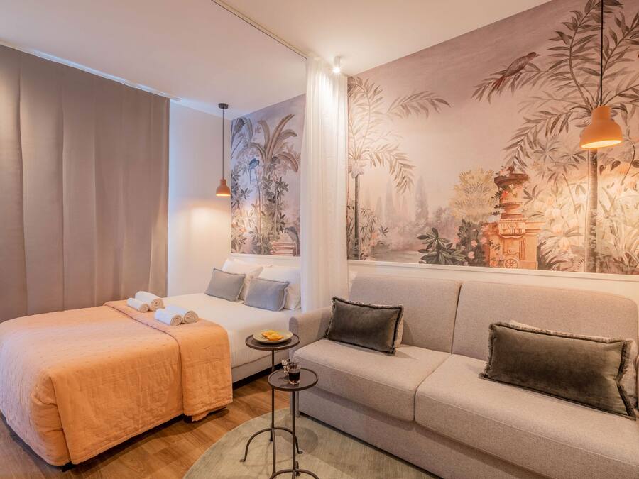 A hotel room with a bed, a sofa, a small table with a drink, and a mural on the wall.