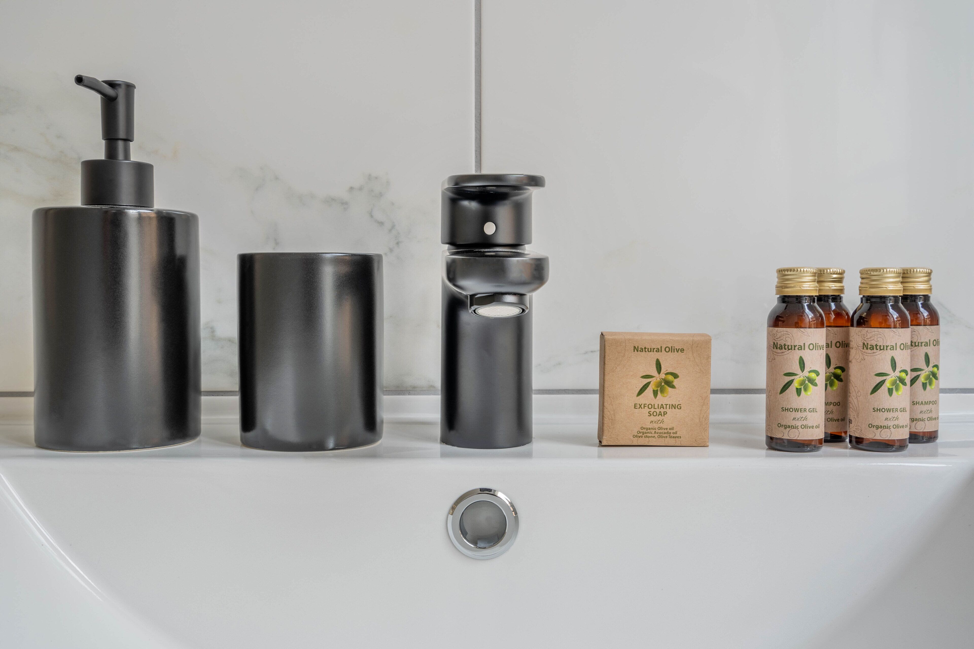 A bathroom with a white sink, a black soap dispenser, a black soap container, a brown box labeled "Natural Olive Exfoliating Soap," and four bottles of "Natural Olive Shower Gel."