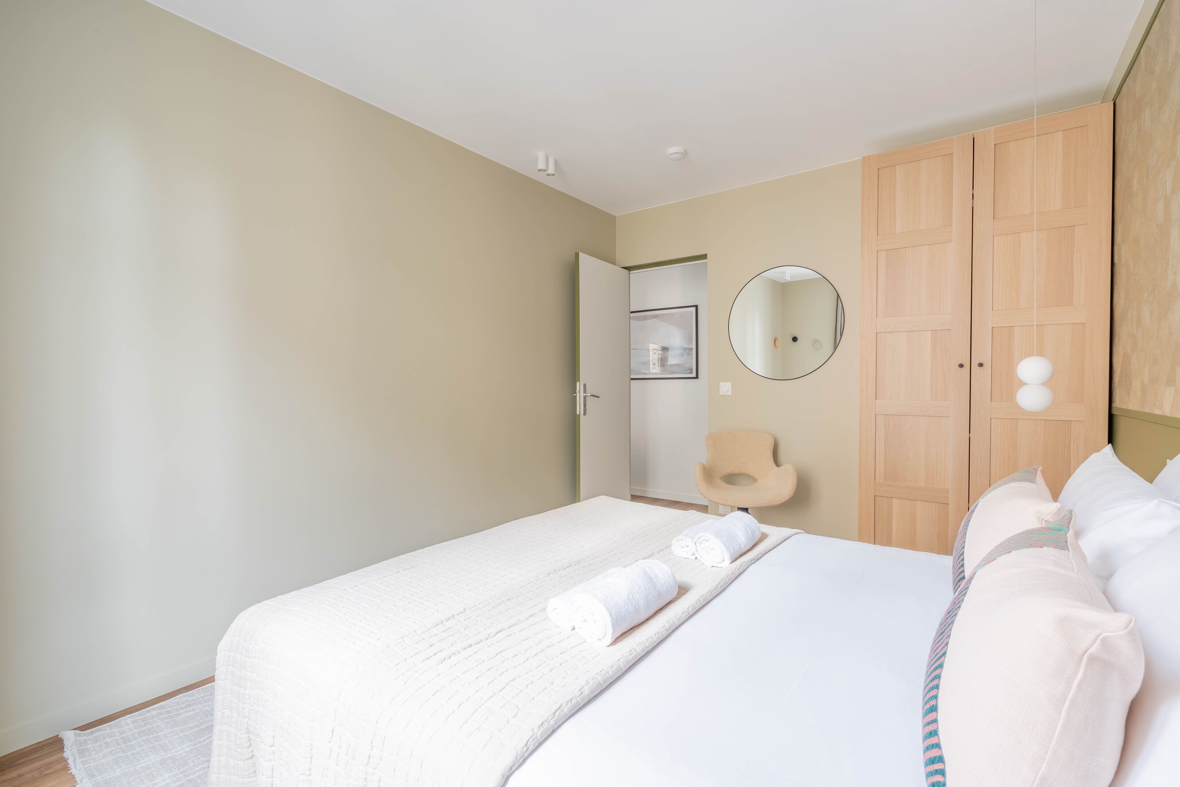 A bedroom with a bed, a wooden wardrobe, a round mirror, and a chair.