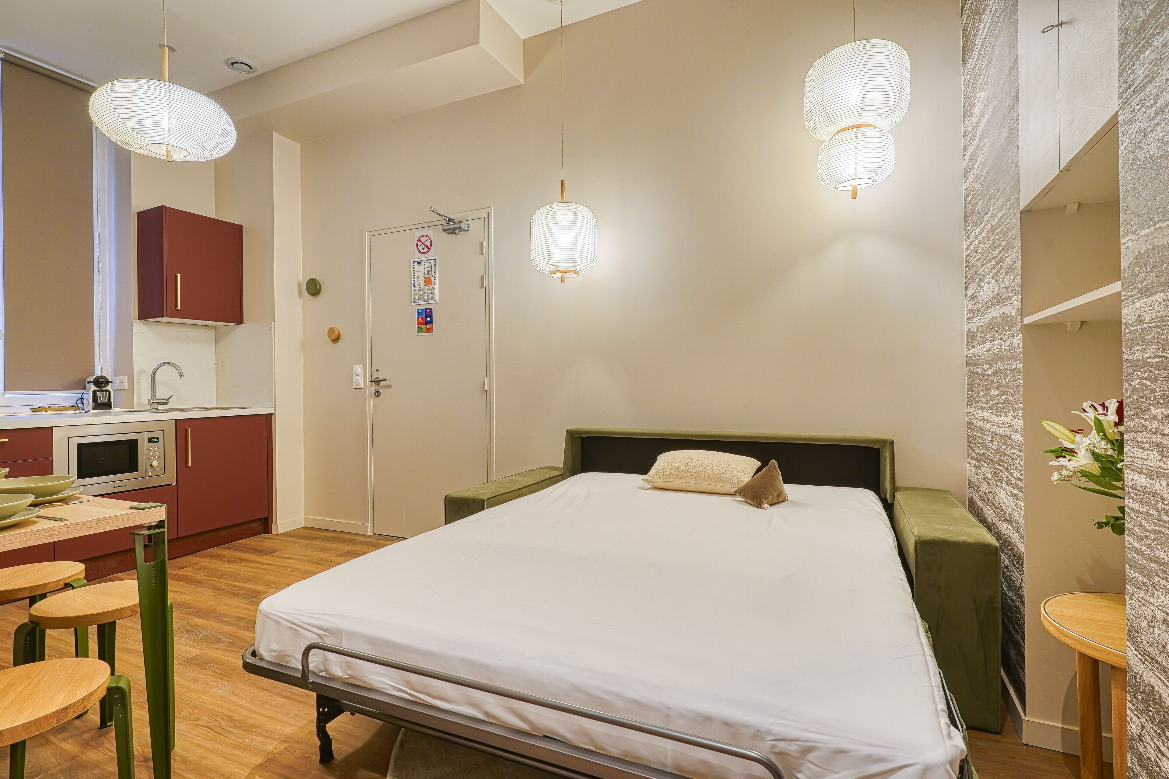 A bedroom with a bed, bedside table, and a small stool. There is a kitchenette with a microwave and cabinets.