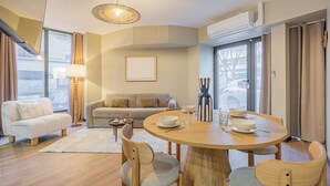 A modern living room with a round wooden dining table, a sofa, and a coffee table. There is a large window with curtains, an air conditioning unit, and a hanging light fixture. - Residence Le Belleville (Paris)