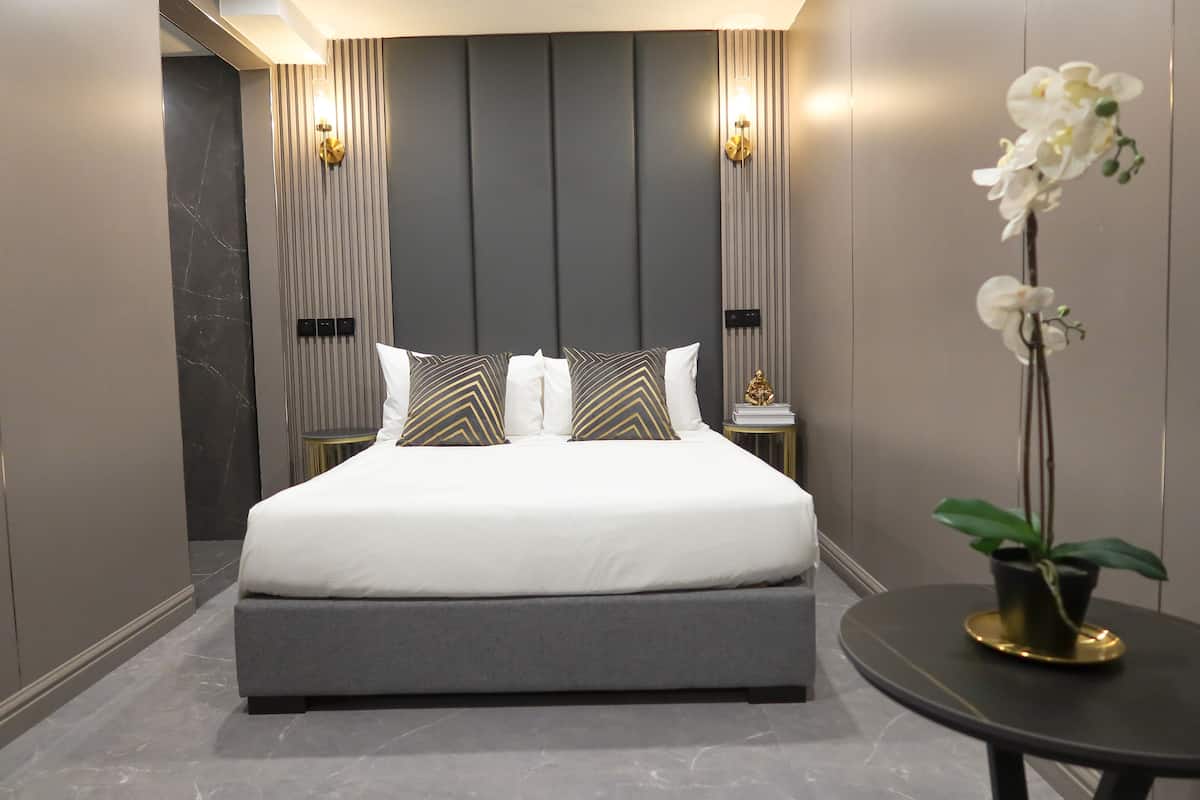 A modern bedroom with a large bed, grey and white bedding, and decorative pillows. There is a small round table with a potted plant to the right.