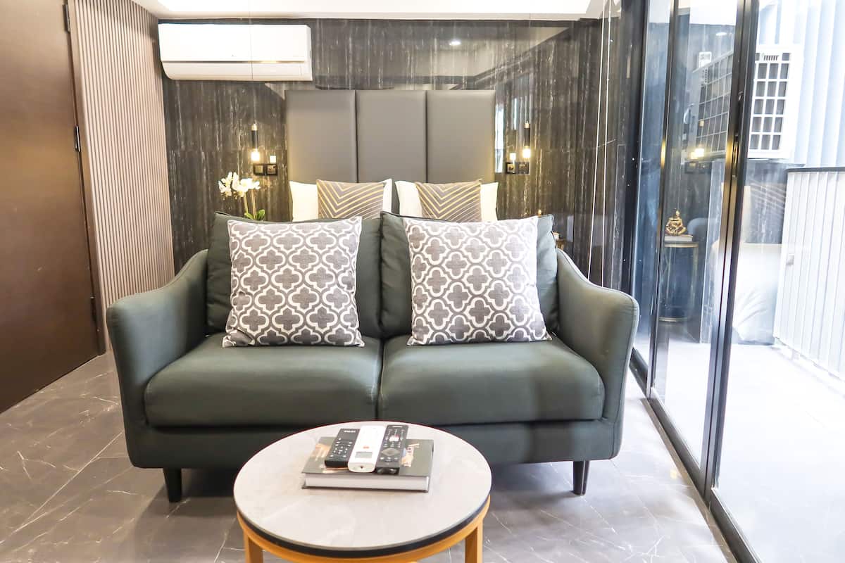 A modern living room with a green sofa, patterned cushions, a small round table with remote controls, and a large window offering a view of the outside.