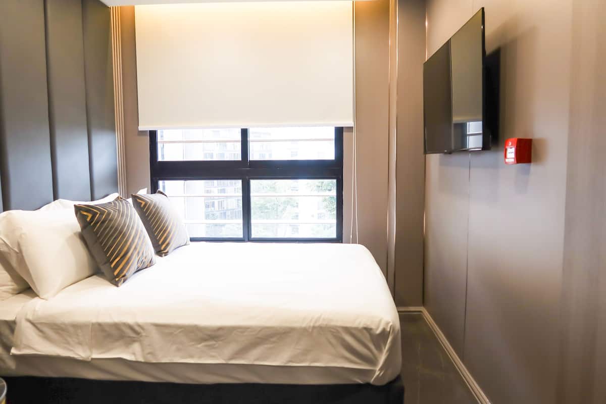 A hotel room with a bed, a window with blinds, a flat-screen TV mounted on the wall, and a red object on the wall.