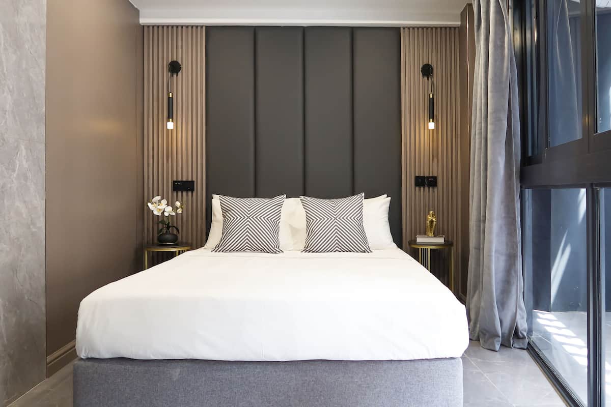 A modern hotel room with a large bed, two bedside tables, a vase with flowers, and a view of the outside through a large window.