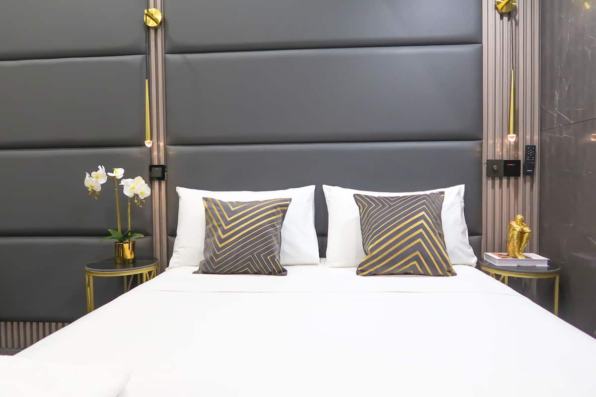 A modern bedroom with a large bed, two decorative pillows, a small side table with a plant, and a wall-mounted control panel.