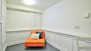 Une petite pièce, bien éclairée, avec un seul canapé orange et une chaise blanche. Les murs sont blancs, et une prise électrique est visible sur le mur.