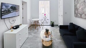 Una sala de estar moderna con un televisor de pantalla plana, una mesa de centro de vidrio, un sofá negro y un comedor con una mesa y sillas blancas.
