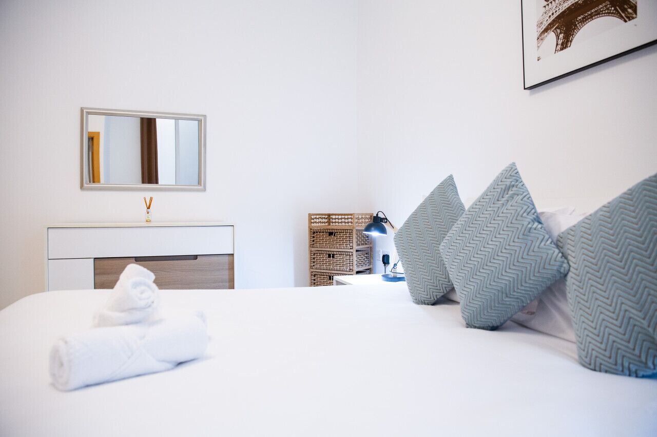 Un letto rifatto con asciugamani bianchi arrotolati, una lampada da comodino e cuscini decorativi.