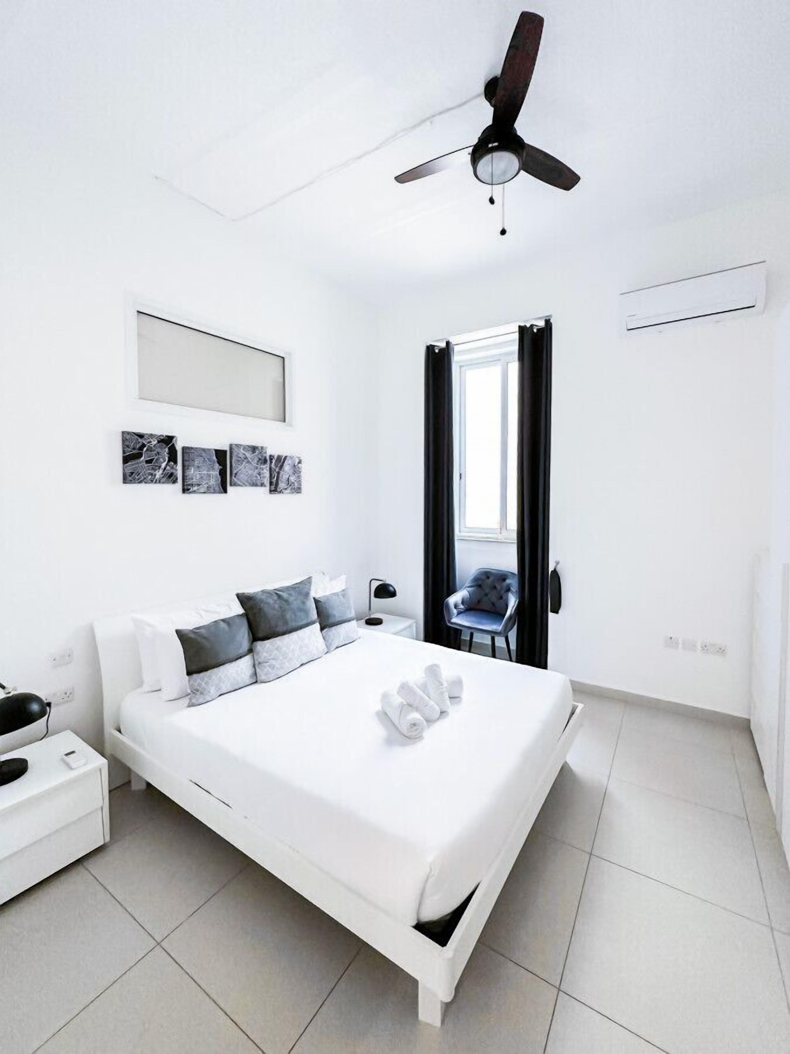Un dormitorio moderno con una cama blanca, ventilador de techo y ventana con cortinas.