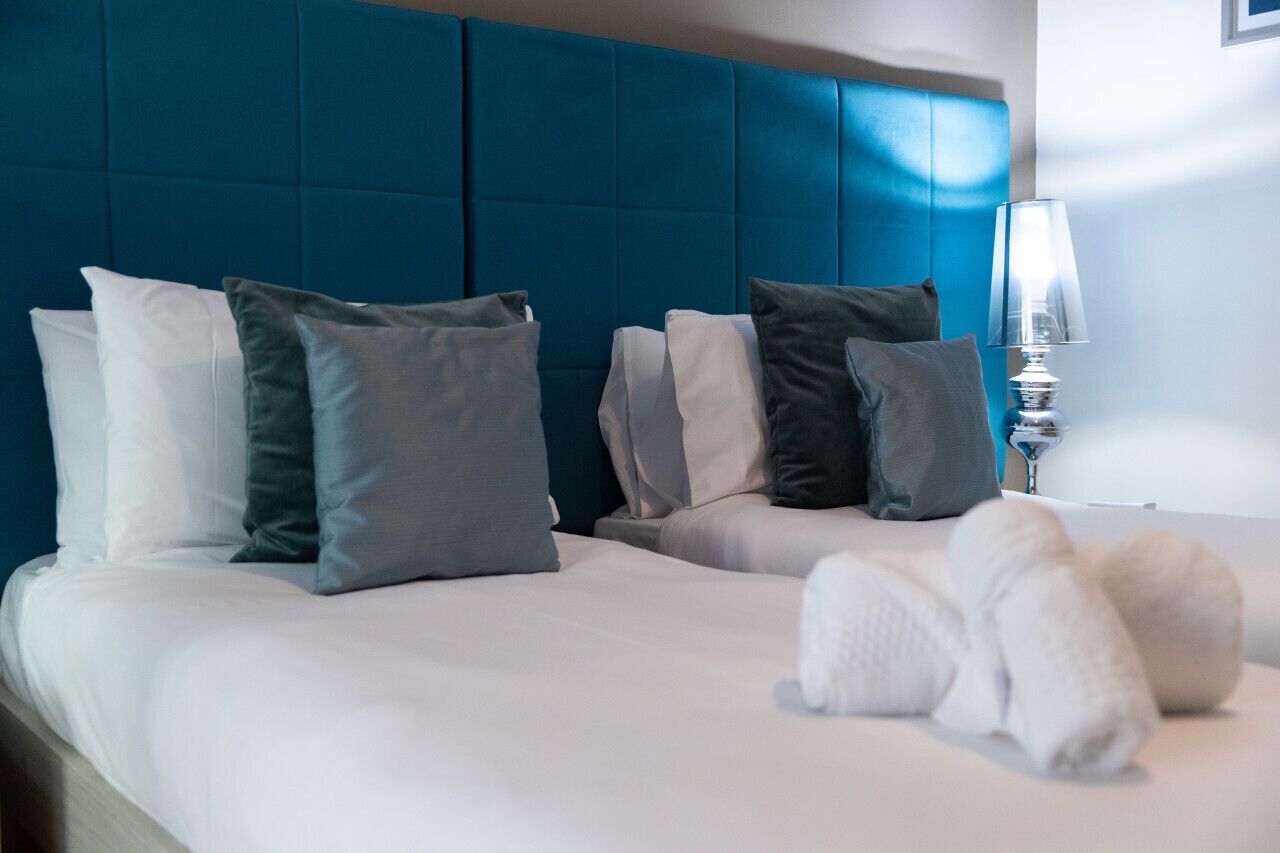 A hotel room with a large blue headboard, multiple pillows, and a white lamp.