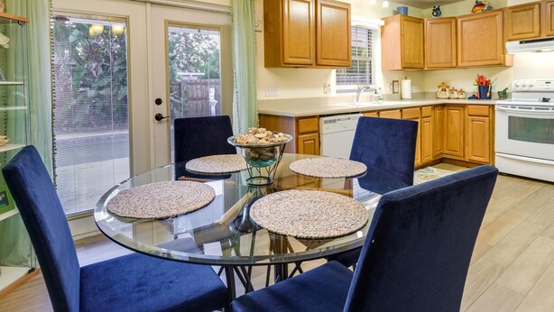 A dining area with a glass table surrounded by four chairs, wooden cabinets, and a kitchen in the background.