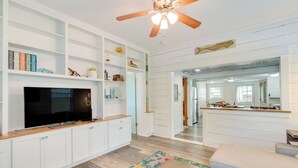 A modern living room with a flat-screen TV, white cabinetry, a ceiling fan, and a kitchen area visible through an open doorway.