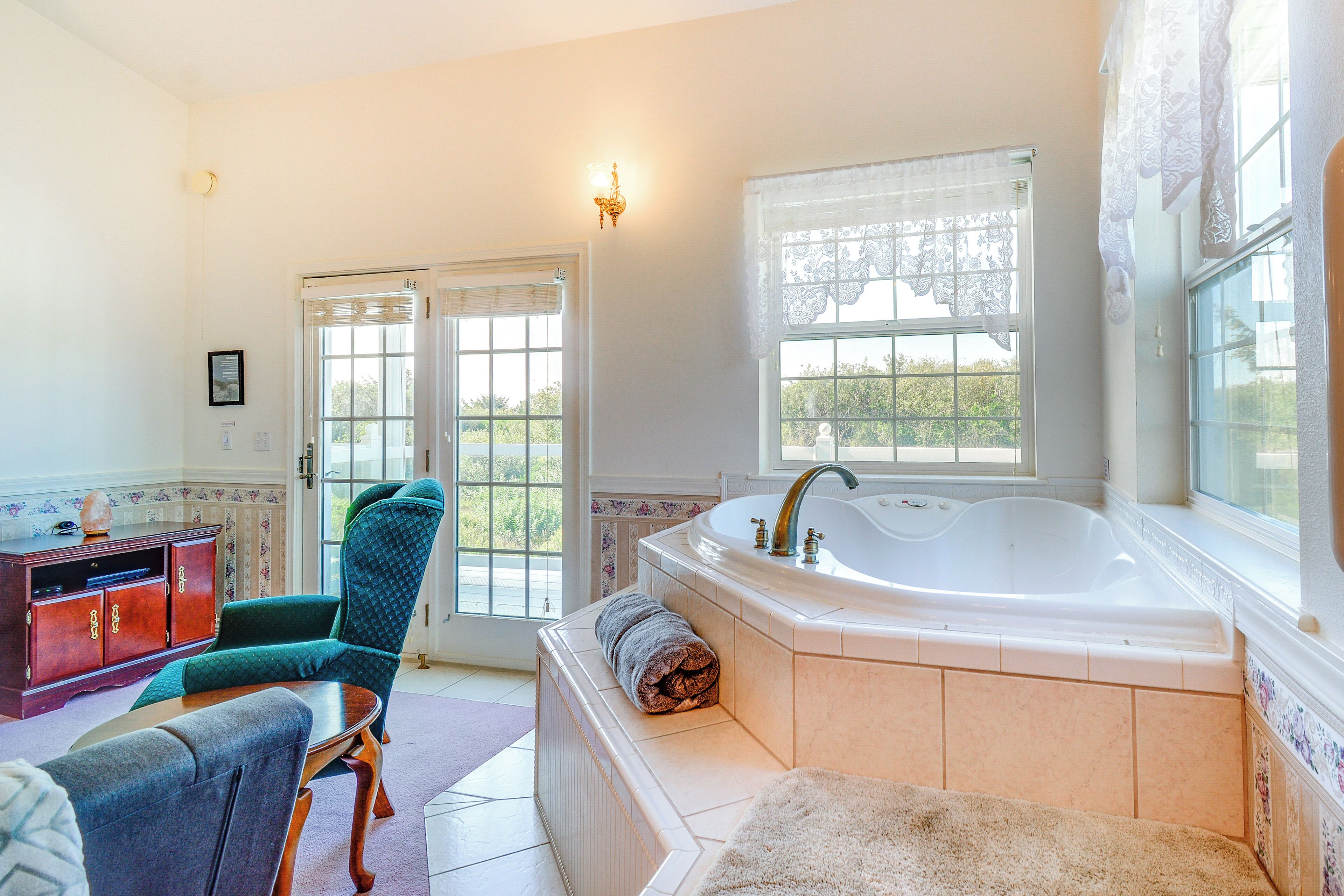 A bathroom with a large bathtub, a wooden cabinet, a green chair, and a window with lace curtains.
