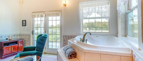 A bathroom with a large bathtub, a wooden cabinet, a green chair, and a window with lace curtains.