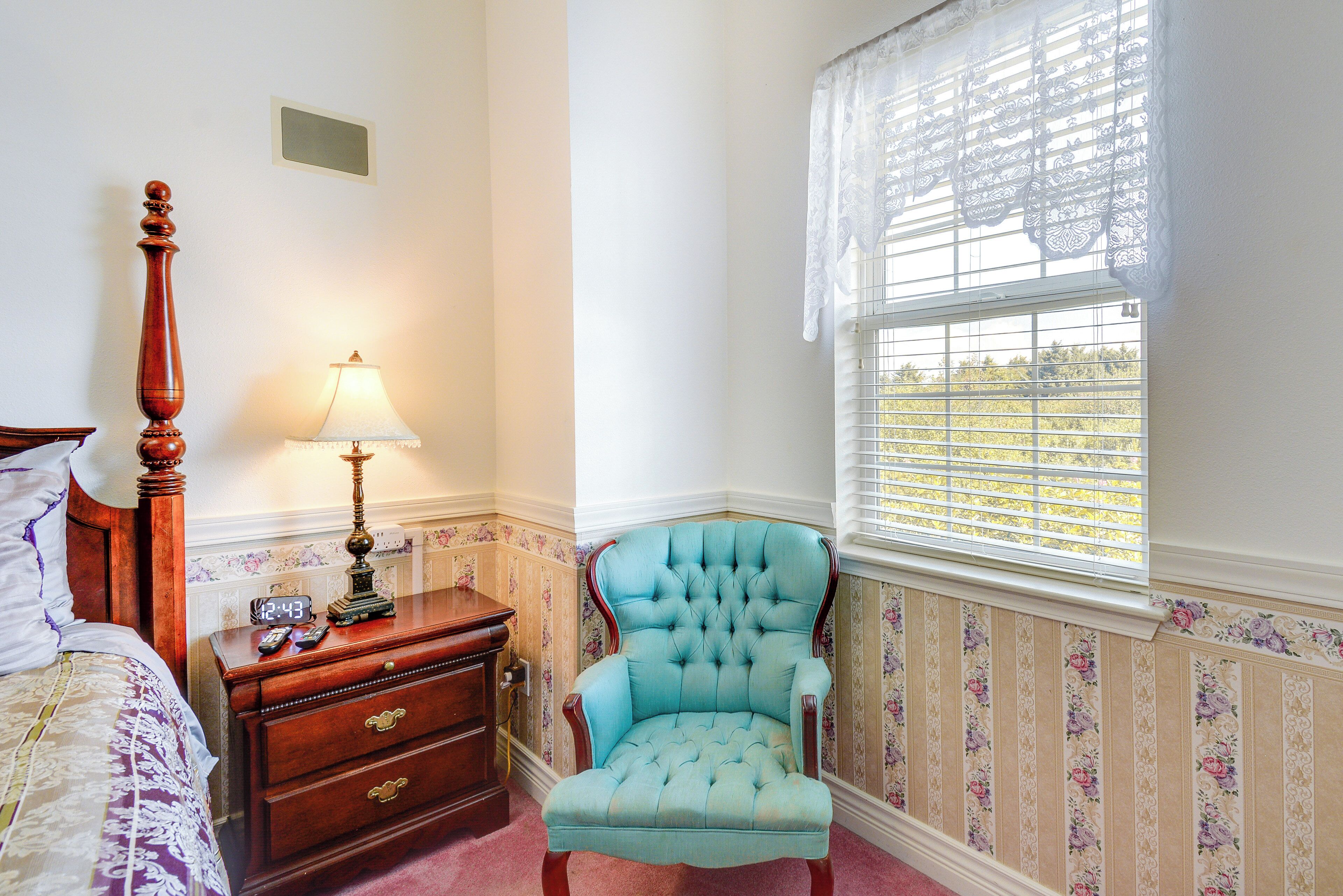 A bedroom with a wooden nightstand, a lamp, a tufted armchair, a bed with a floral bedspread, and a window with lace curtains.