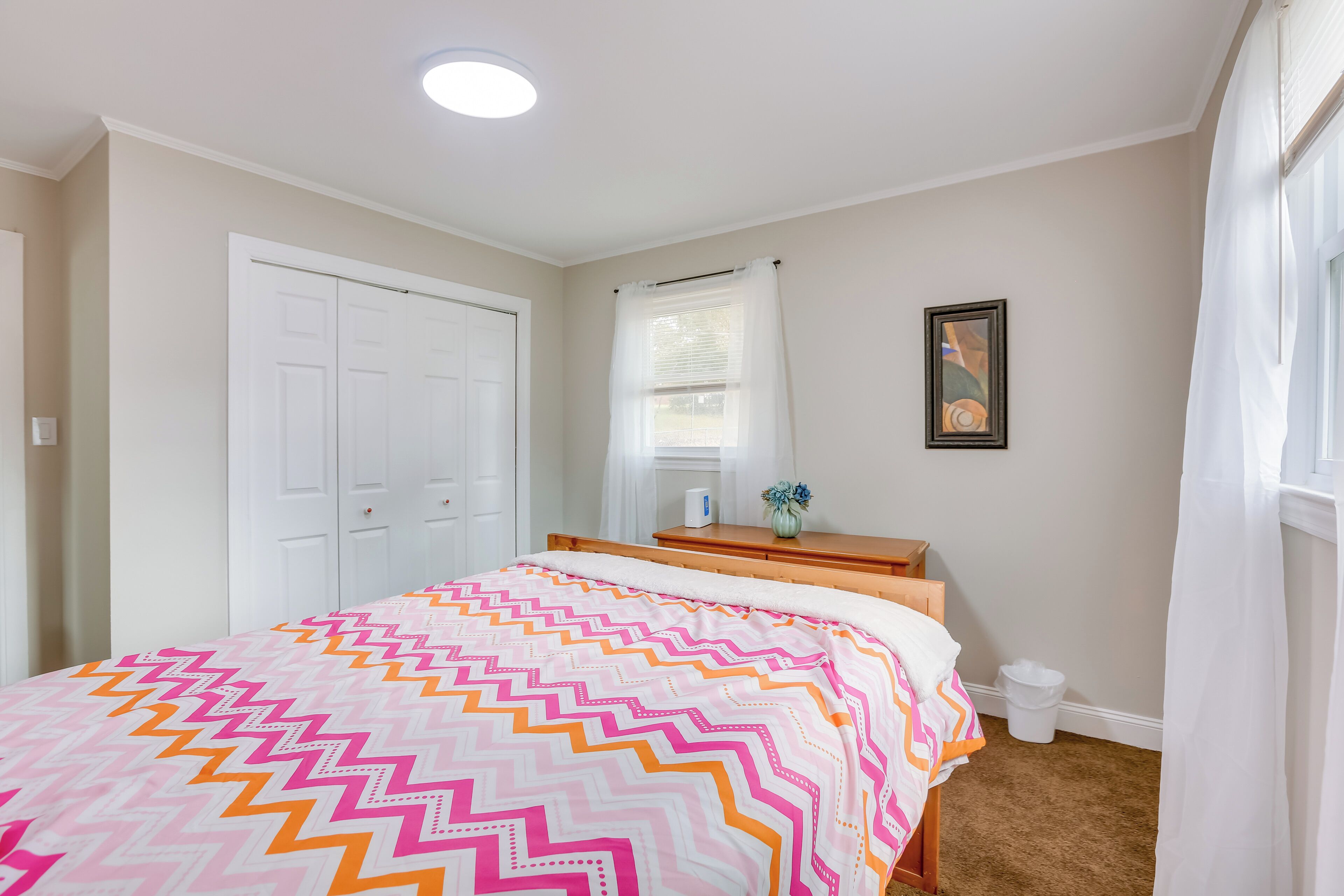 A bedroom with a bed, a dresser, a window with curtains, a framed picture on the wall, and a white door.