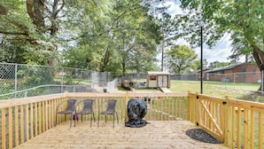 Terraza de madera con sillas y una parrilla cubierta, rodeada por una valla y árboles.