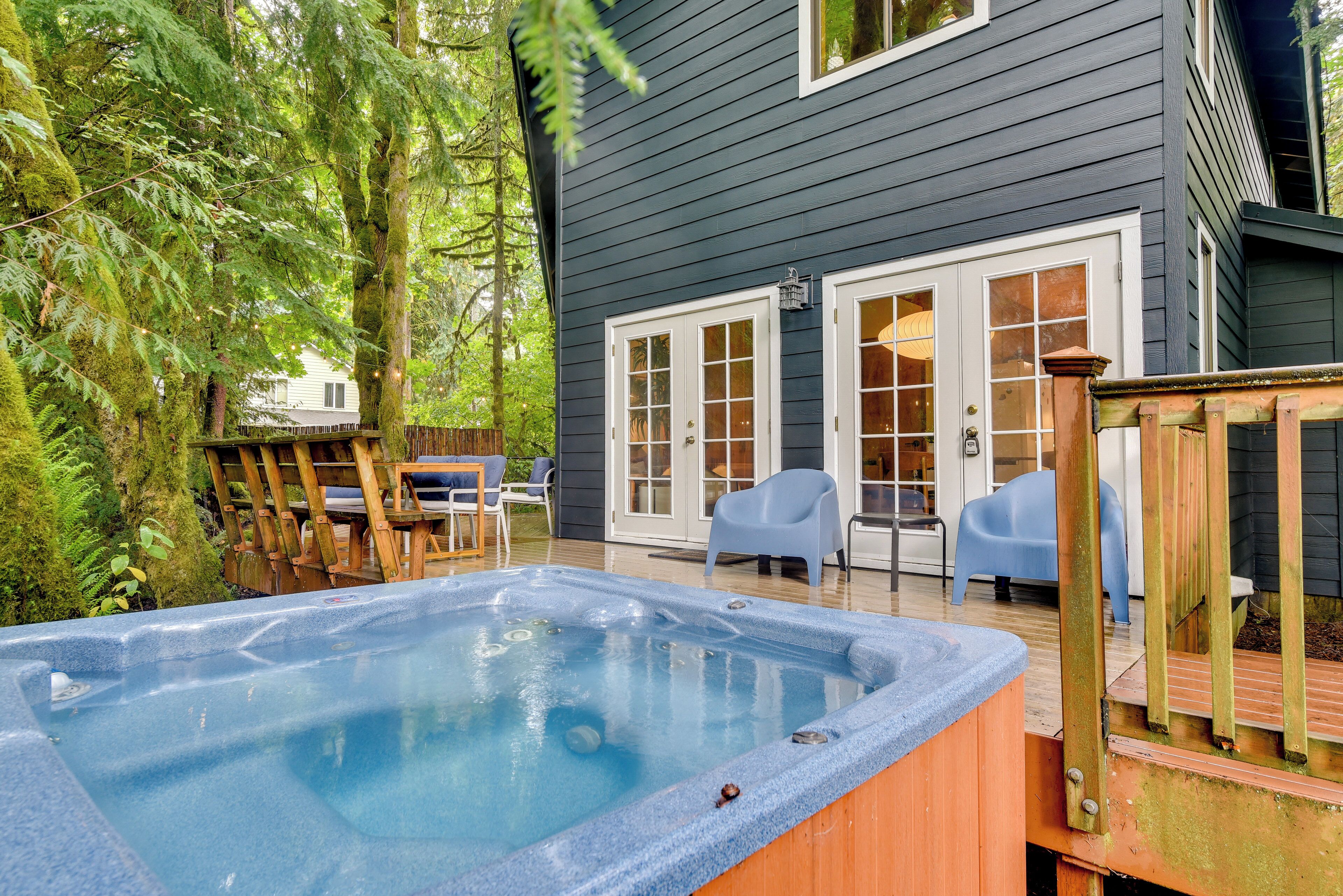 A hot tub in a backyard with a wooden deck, a dining area with chairs, and a house with dark siding and white doors.