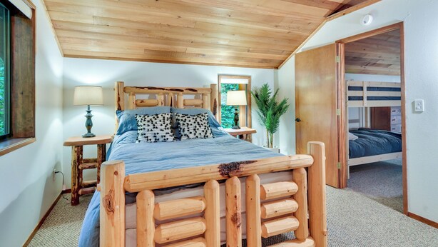 A wooden-framed bed with a blue bedspread, a nightstand with a lamp, and a window with a view of greenery. - Rhododendron Creekside Cabin w/ Hot Tub & Fire Pit (Rhododendron)