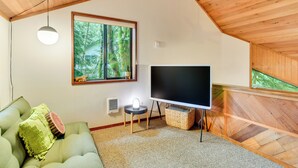 A cozy living room with a green sofa, a flat-screen TV, a small side table, and a window overlooking a forest. - Rhododendron Creekside Cabin w/ Hot Tub & Fire Pit (Rhododendron)