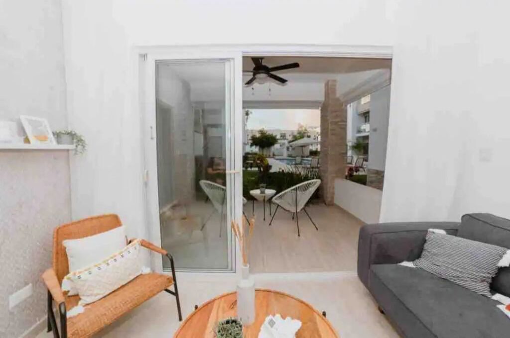 Ein modernes Wohnzimmer mit einem grauen Sofa, einem Holztisch und einem Korbstuhl. Eine Glastür führt zu einer Außenpatio mit einem Deckenventilator, einem Tisch und Stühlen.
