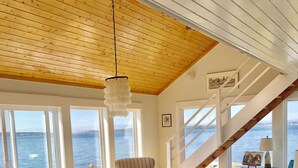 Een ruime woonkamer met uitzicht op het water, een houten plafond en een trap naar een balkon.