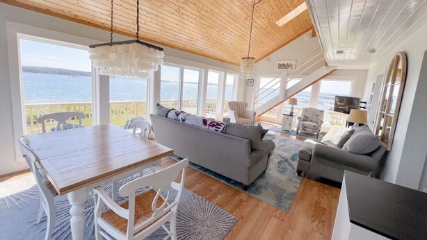 Een ruime woonkamer met een houten plafond, grote ramen met uitzicht op zee, een eettafel met stoelen en een bank.