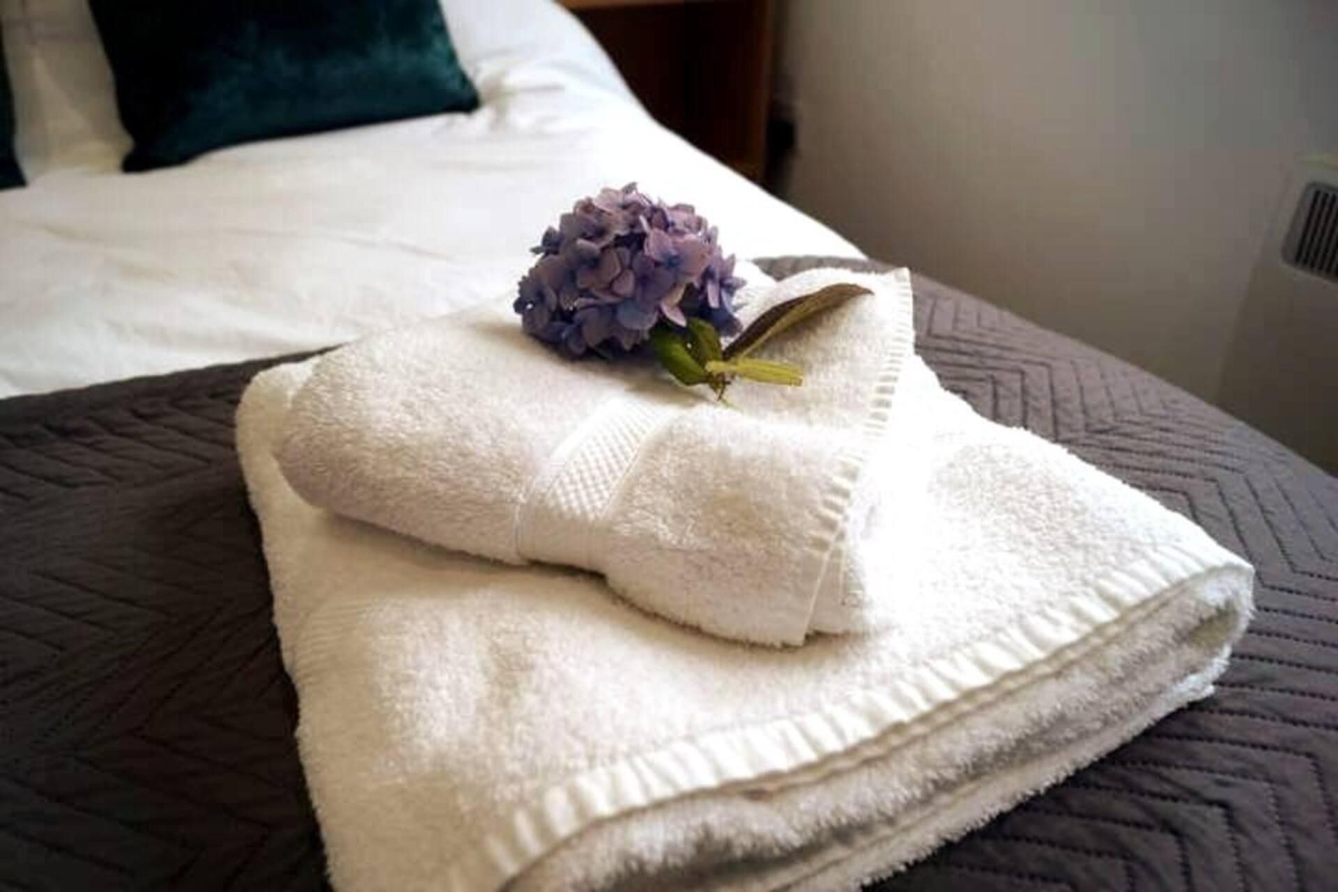 Una toalla blanca doblada con un detalle de flor morada sobre una cama con cubierta acolchada marrón oscuro.