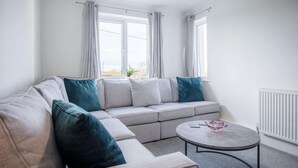 A modern living room with a grey sectional sofa, teal cushions, a round coffee table, and large windows with curtains.