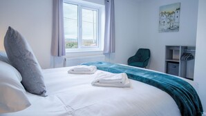 A neatly made bed with white linens and a teal blanket, a grey pillow, and two folded towels on the bed. There is a window with curtains, a chair, and a painting on the wall.