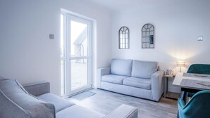 A modern living room with a grey sofa, a matching armchair, a teal armchair, and a small table with a lamp. There are two windows with decorative frames and a door leading outside.