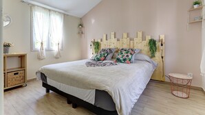 Una camera da letto con un letto, una testiera in legno, un tavolino rotondo, uno scaffale con piante e una finestra con tende.
