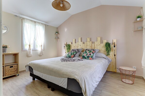 A bedroom with a bed, a wooden headboard, a small round table, a shelf with plants, and a window with curtains.