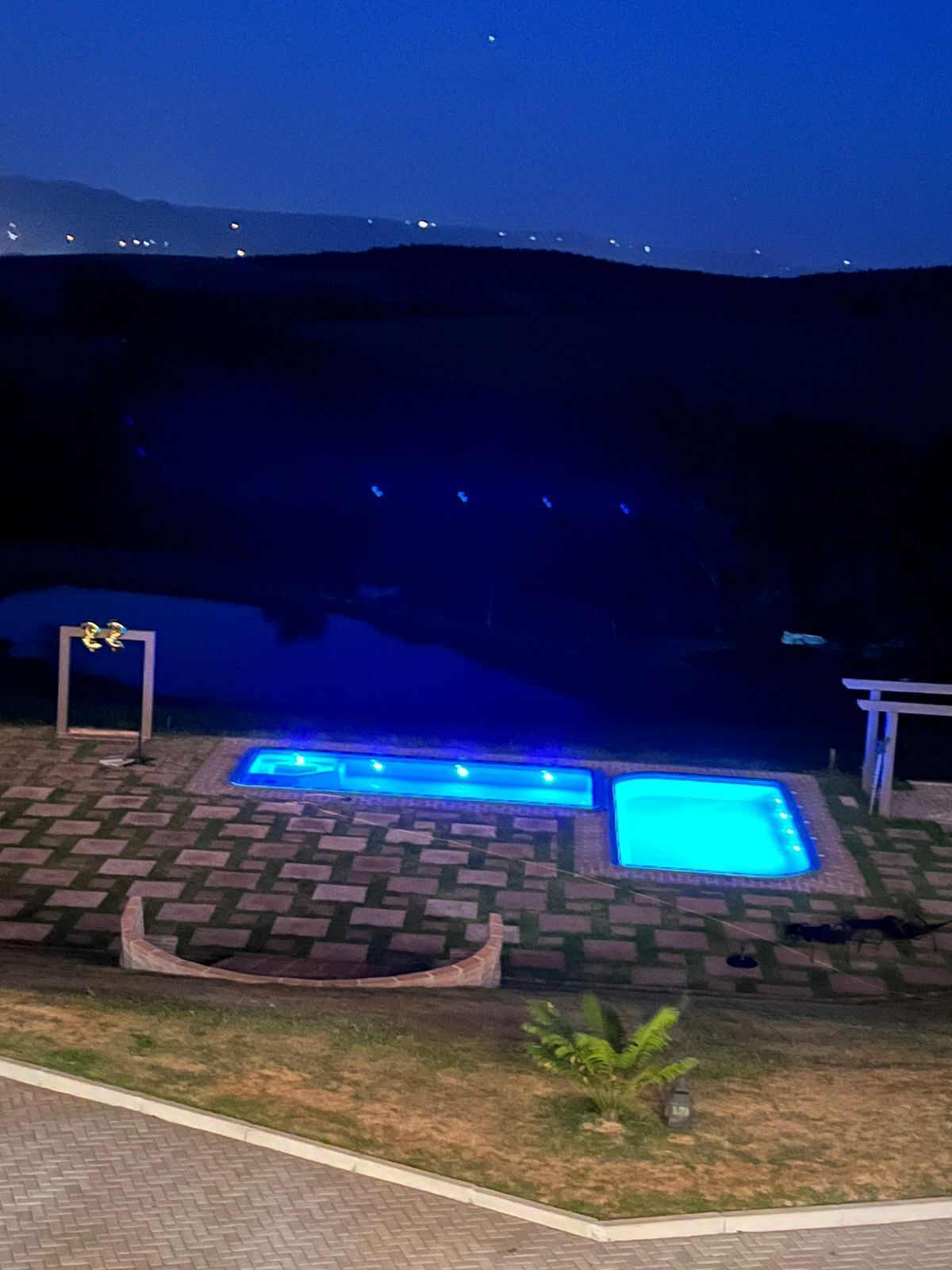 Área de piscina com um caminho de tijolos que a leva, cercada por grama e uma pequena planta.