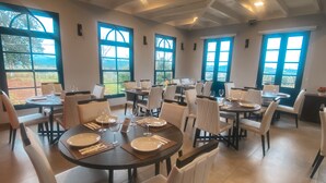 Un espace repas avec des tables rondes, des chaises blanches et de grandes fenêtres donnant sur l’extérieur.