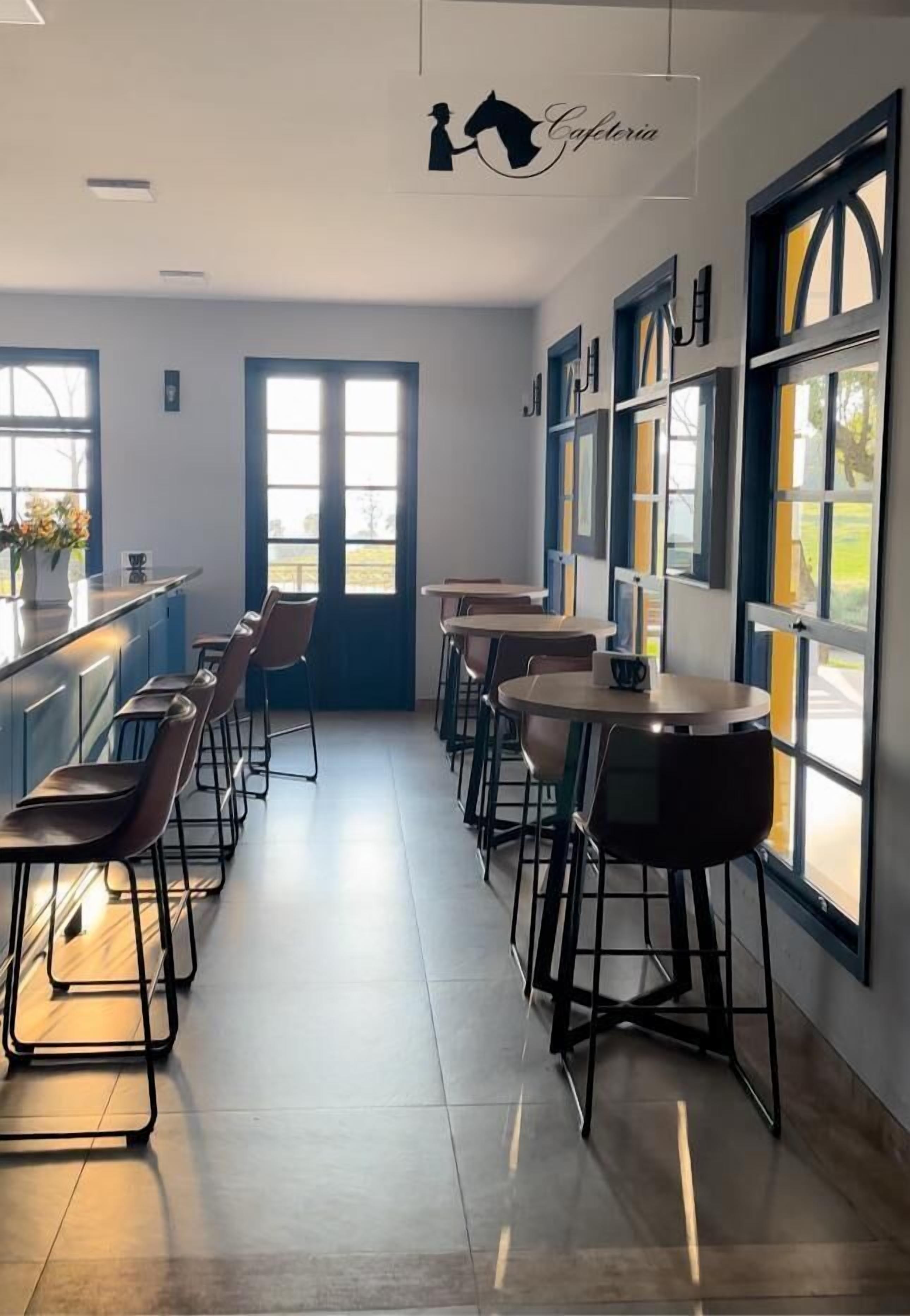Um café com um balcão comprido, banquetas e mesas. Há grandes janelas com vista para o exterior.