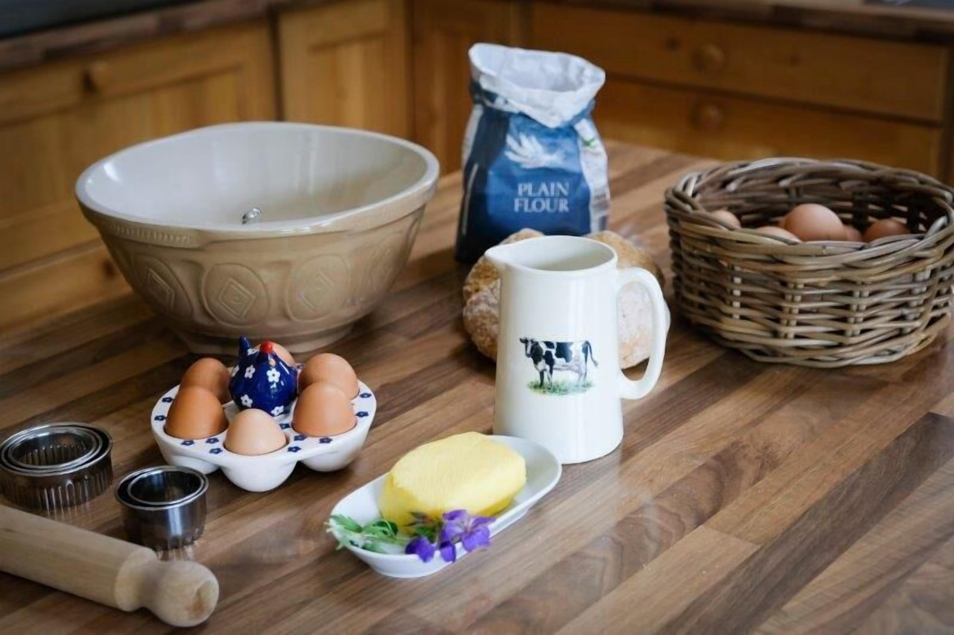 A kitchen counter with a large bowl, a jug of plain flour, a basket of eggs, a mug with a cow design, a plate with butter, and a rolling pin.