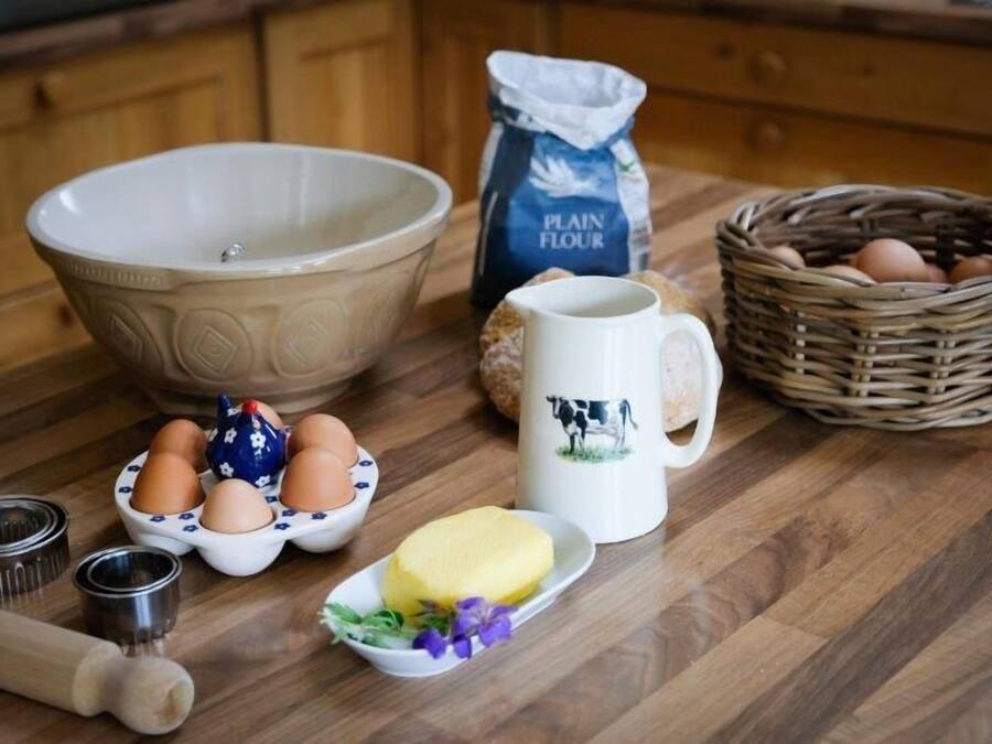 A kitchen counter with a large bowl, a jug of plain flour, a basket of eggs, a mug with a cow design, a plate with butter, and a rolling pin.