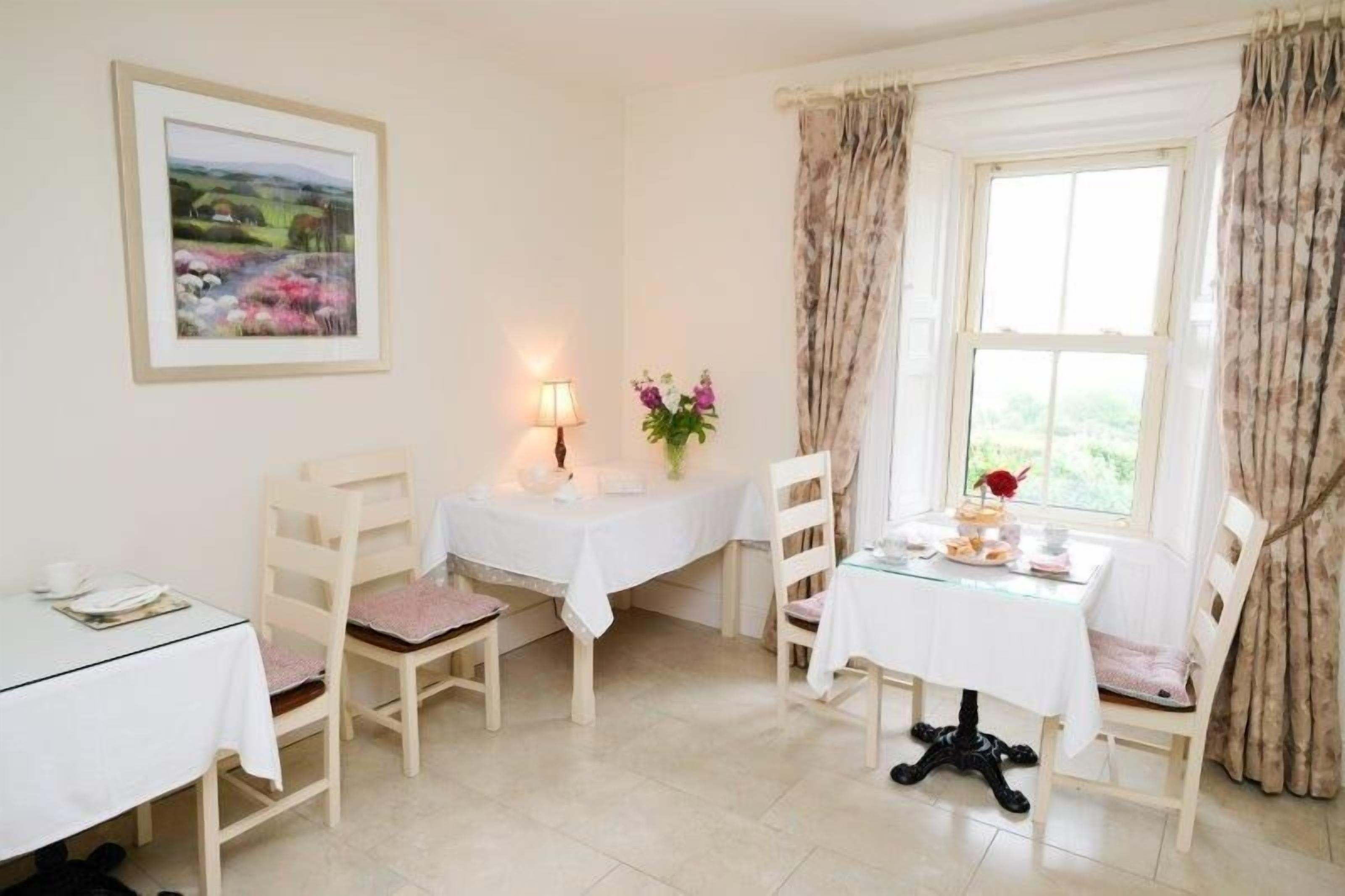 A dining area with two tables, each with a white tablecloth and chairs. There is a framed picture on the wall, a lamp on one table, and a vase with flowers on the other.