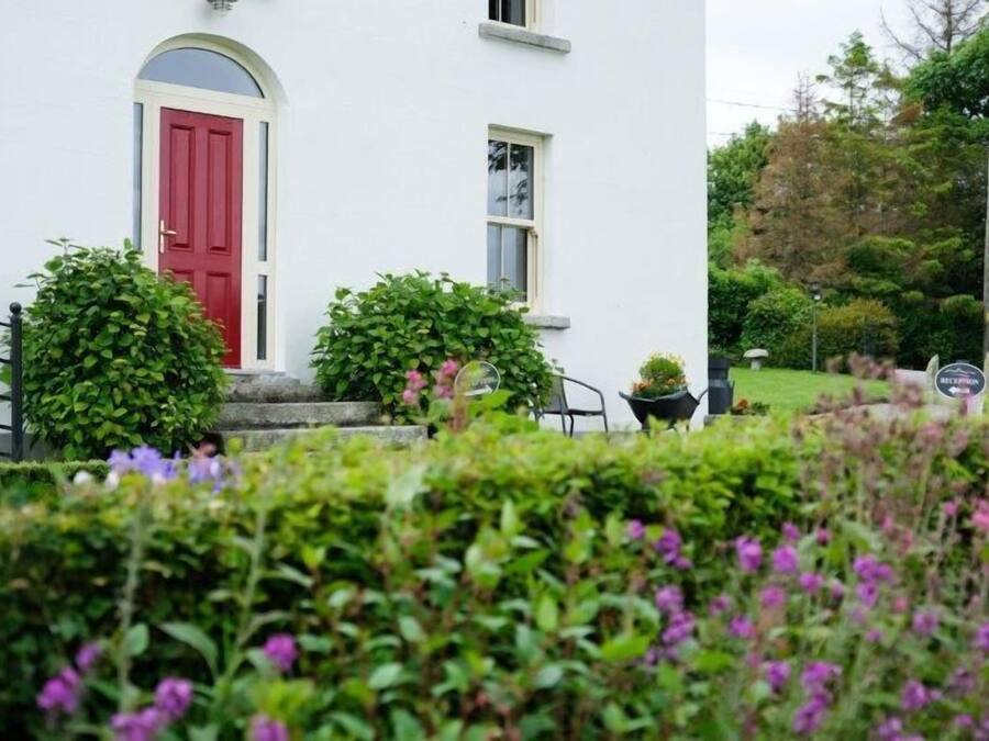 A white building with a red door, surrounded by lush greenery and purple flowers.
