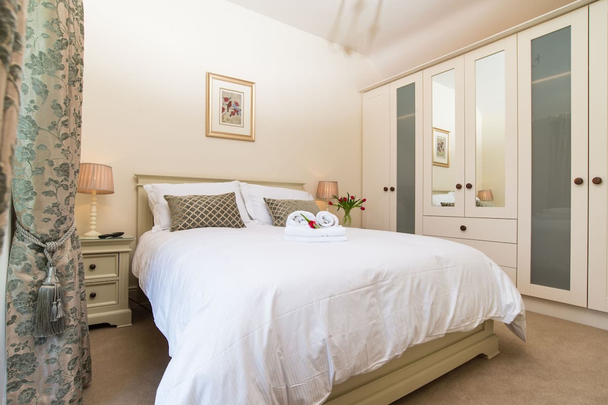 A neatly made bed with a white comforter and pillows, a bedside table with a lamp, and a wardrobe with glass doors.